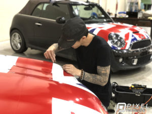A man installing a custom-printed vinyl vehicle wrap of the United Kingdom Union Jack flag on two Cooper Mini cars.